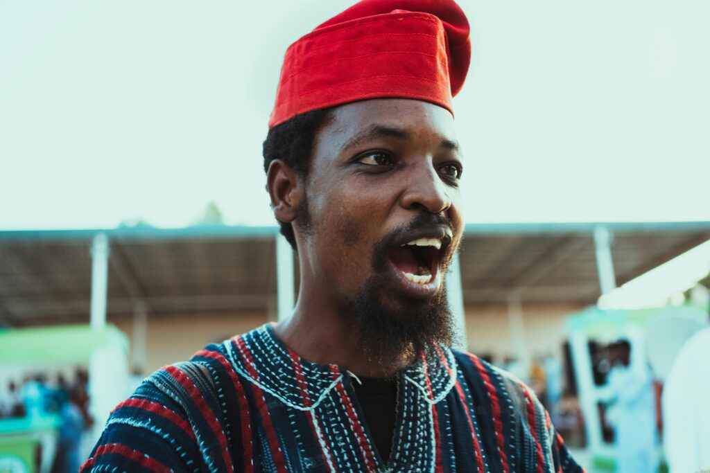 A joyful Nigerian man wearing a traditional red cap during an outdoor cultural event.